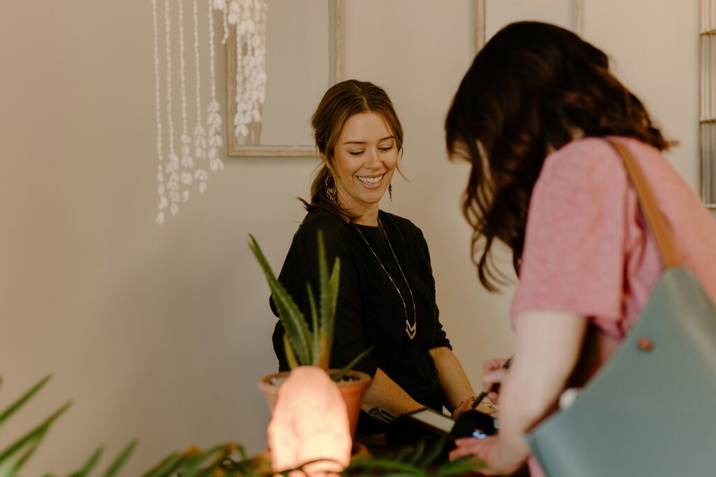 Uma recepcionista ou lojista sorridente, com cabelo castanho apanhado e uma camisola preta, observa uma cliente de costas enquanto esta utiliza um terminal de pagamento no balcão. No balcão estão uma planta de aloé vera e uma lâmpada de sal rosa acesa.