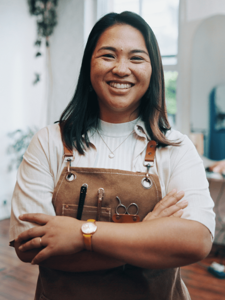 Retrato de uma profissional de salão sorridente de cabelo escuro, a olhar diretamente para a câmara com os braços cruzados. Usa uma camisa clara sob um avental de lona castanho equipado com ganchos de cabeleireiro e tesoura.