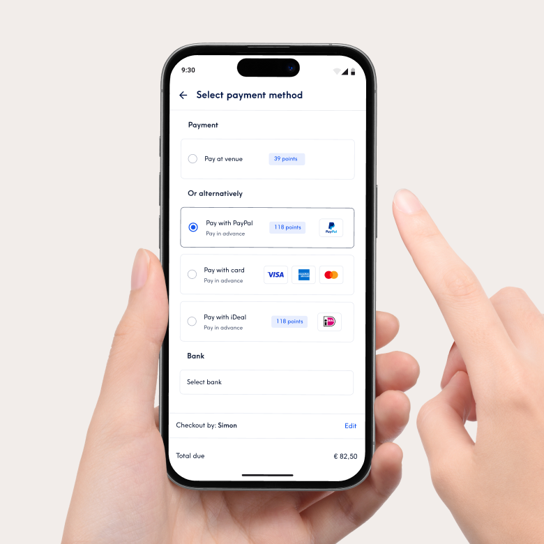 Primeiro plano de um smartphone que mostra a interface de pagamento da aplicação Treatwell para selecionar o método de pagamento. As opções incluem o pagamento no local ou o pagamento antecipado com PayPal, cartão ou iDeal. Uma mão segura o telemóvel e um dedo aponta para o ecrã.