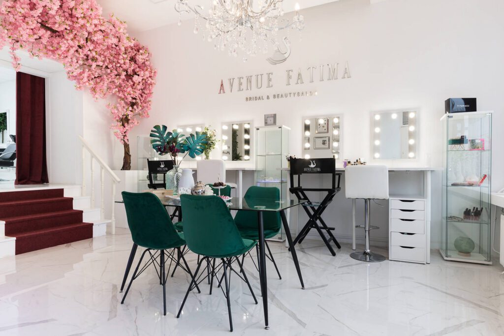 Um salão de beleza e de noivas luminoso e elegante chamado "Avenue Fatima". O espaço dispõe de várias bancadas de maquilhagem com espelhos iluminados, cadeiras de veludo verde-esmeralda em redor de uma mesa de vidro e uma cerejeira rosa decorativa.