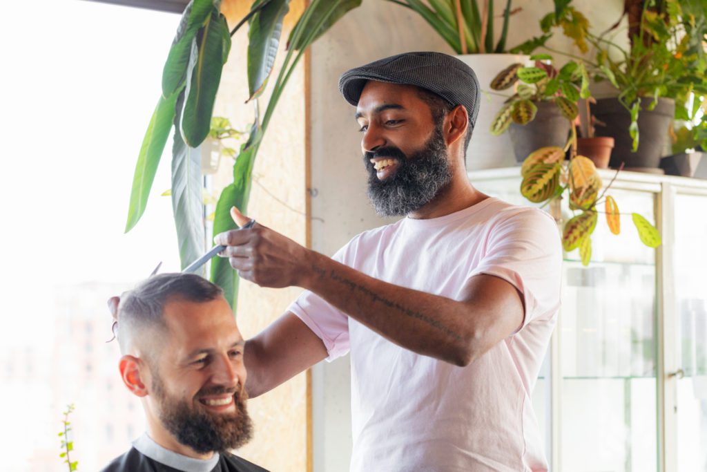Um barbeiro sorridente com barba escura e uma boina cinzenta utiliza tesoura e pente no cabelo de um cliente sorridente num salão bem iluminado e decorado com plantas verdes.