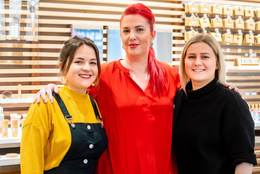 Três profissionais de salão sorridentes posam juntas abraçadas em frente a uma parede de ripas de madeira que exibe produtos cosméticos. A mulher do centro tem o cabelo vermelho vivo e uma blusa vermelha.