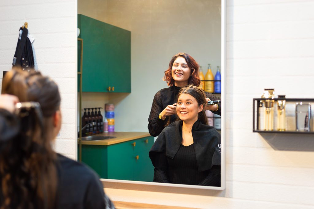 Uma cabeleireira sorridente de cabelo acaju modela o cabelo de uma cliente sorridente com uma placa de alisar, vista através do reflexo de um grande espelho retangular num salão moderno com móveis verde-esmeralda.