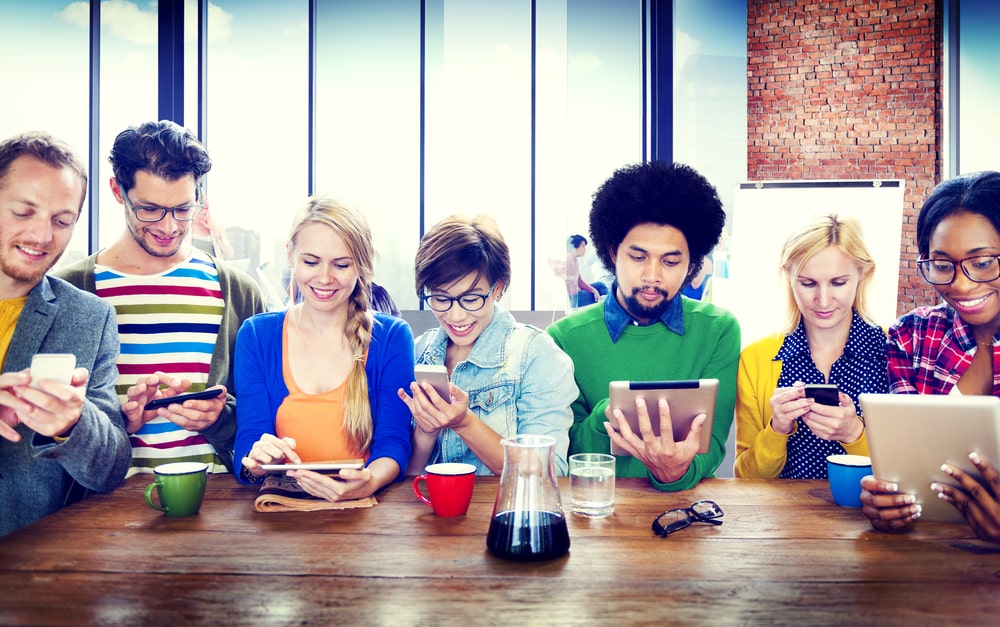 Grupo diverso de pessoas a uma mesa a usar smartphones e tablets, simbolizando a formação online de cabeleireiros e barbeiros