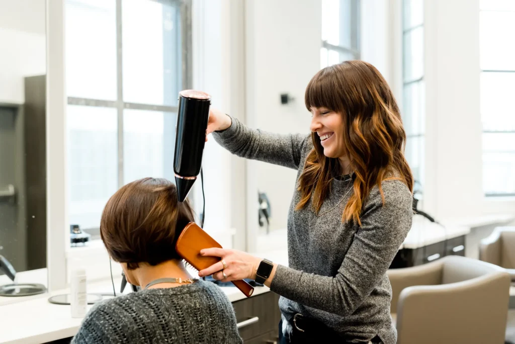 Uma cabeleireira sorridente faz brushing e escova o cabelo de um cliente num salão luminoso e moderno.