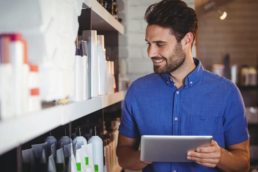 Um dono de salão sorridente de camisa azul a olhar para um tablet junto a prateleiras de produtos de cabelo e beleza.