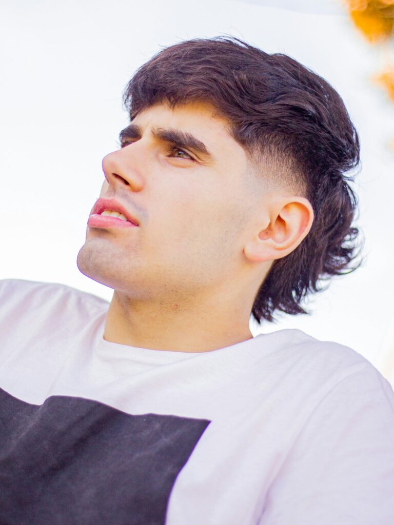 [ALT_TEXT: A young man with a sharp fade mullet haircut wearing a white t-shirt sitting by a road on a bright day.]