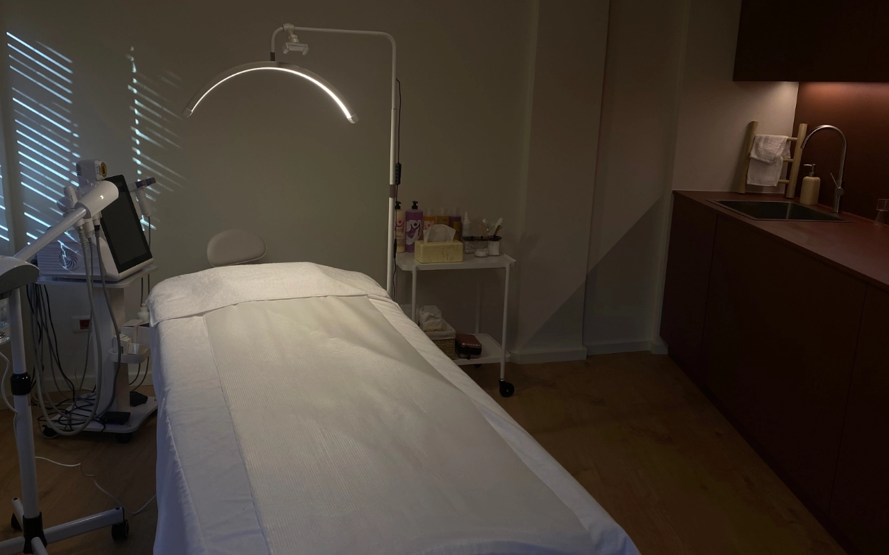 Uma sala de tratamento focada com uma cama branca sob um candeeiro LED em arco, equipamento profissional de estética com ecrã digital, produtos de cuidados de pele num carrinho e armários de madeira escura com lavatório ao fundo.