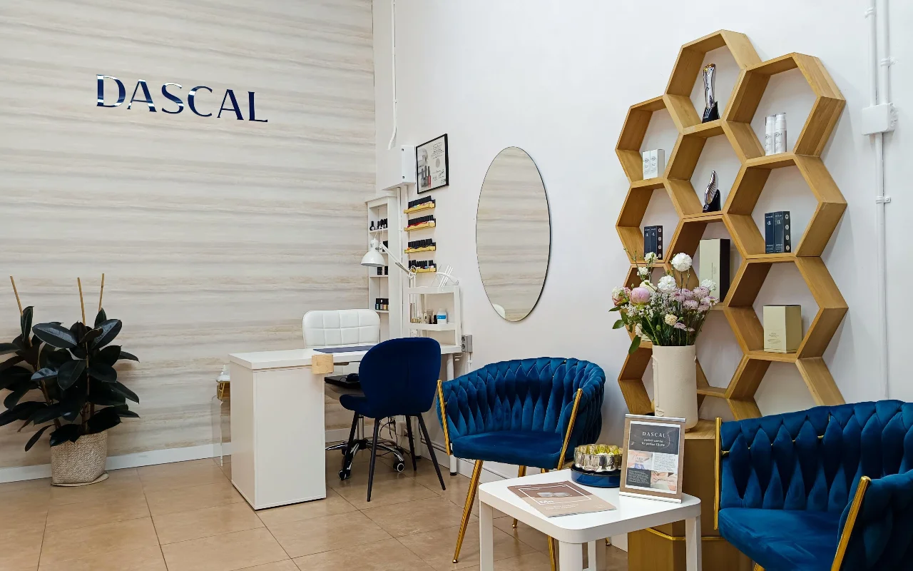 Um interior elegante de salo de beleza com cadeiras de veludo azul profundo e uma estante hexagonal de madeira na parede. Uma mesa de manicure branca est no canto, contra uma parede de textura clara com a palavra "DASCAL" em letras azuis.