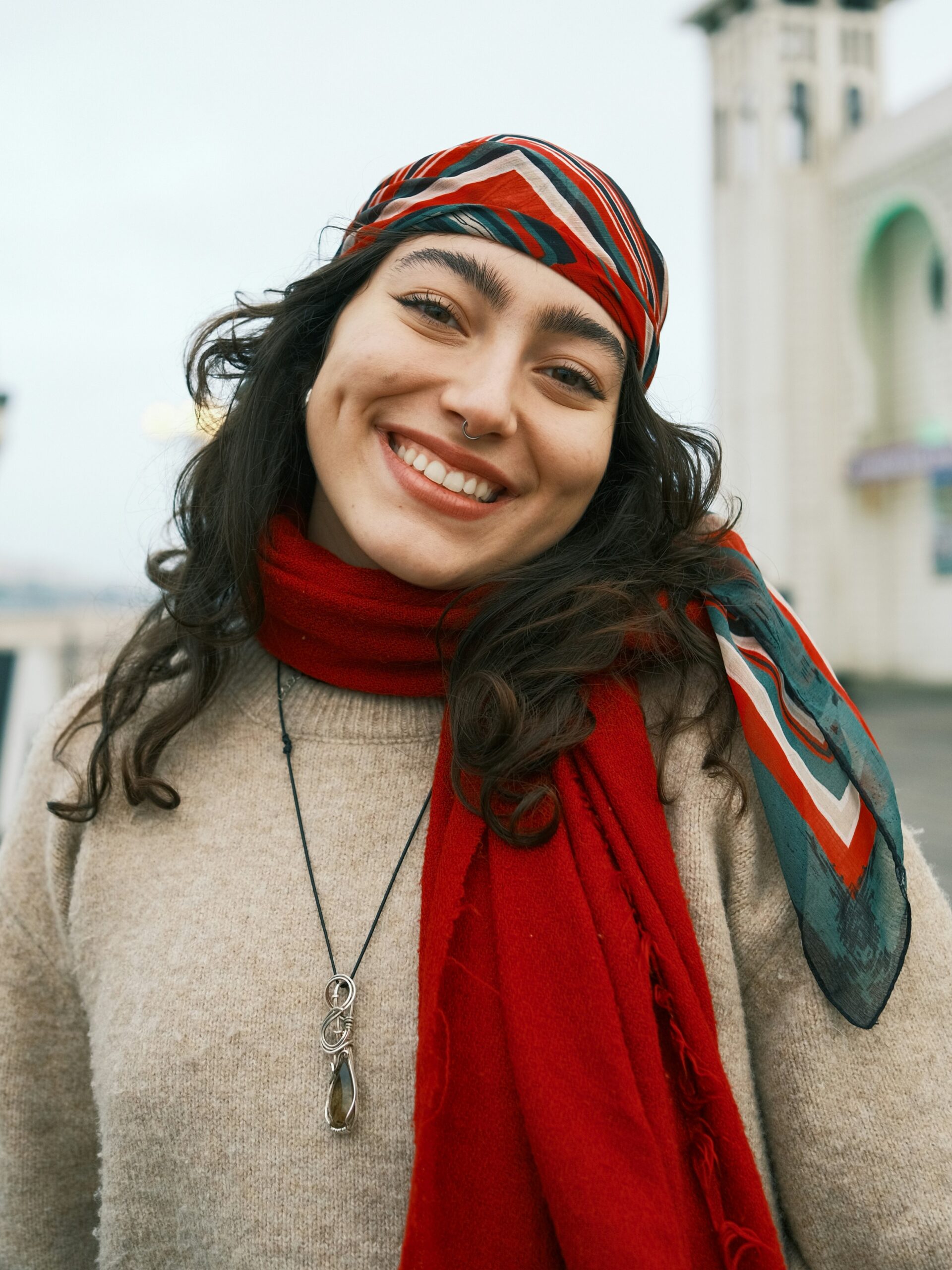 Mulher sorridente ao ar livre num cais, com cabelo escuro comprido, um lenço às riscas vermelho, branco e verde na cabeça e um cachecol vermelho ao pescoço