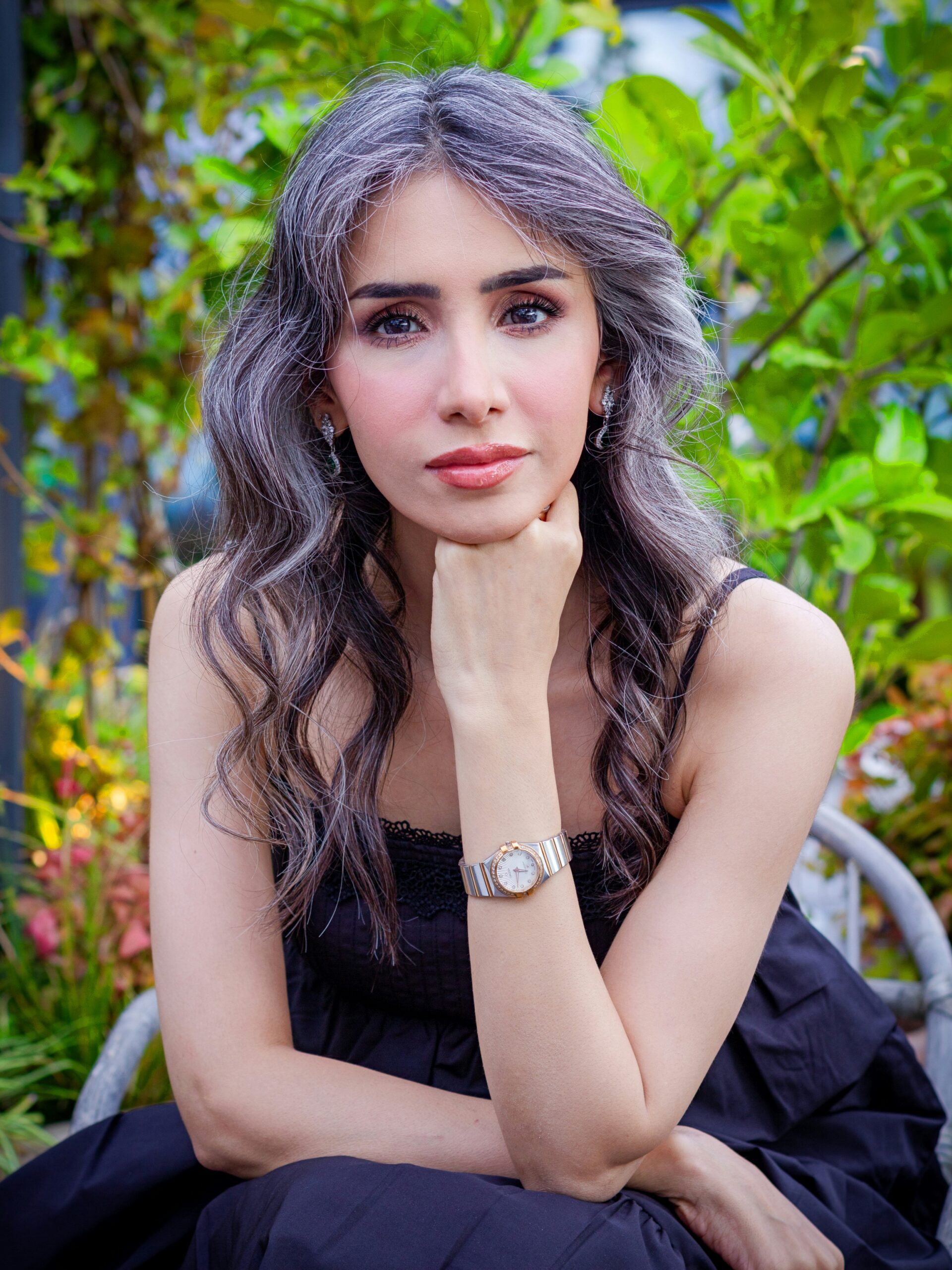 Mulher com cabelo comprido, ondulado, escuro e com grisalhos disfarçados, sentada numa cadeira de jardim a posar com a mão no queixo