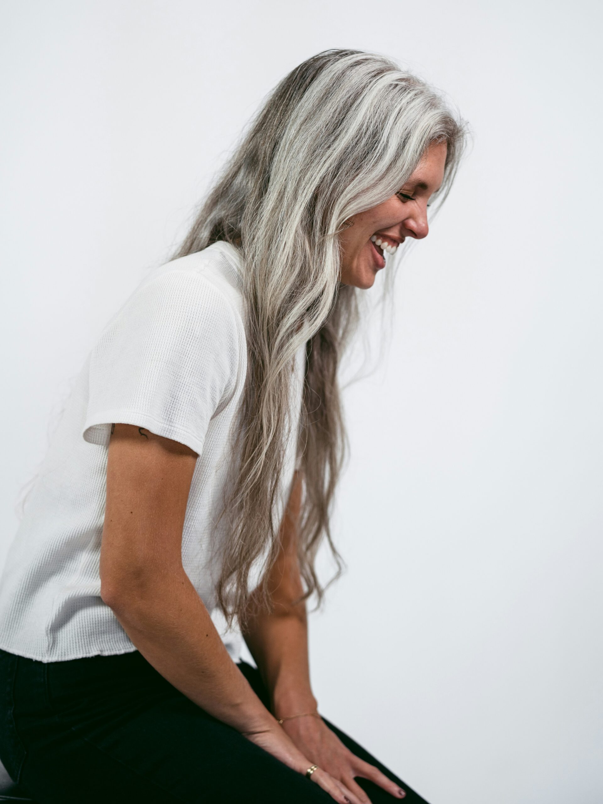 Vista lateral de uma mulher sentada num banco, a rir, com cabelo grisalho muito comprido