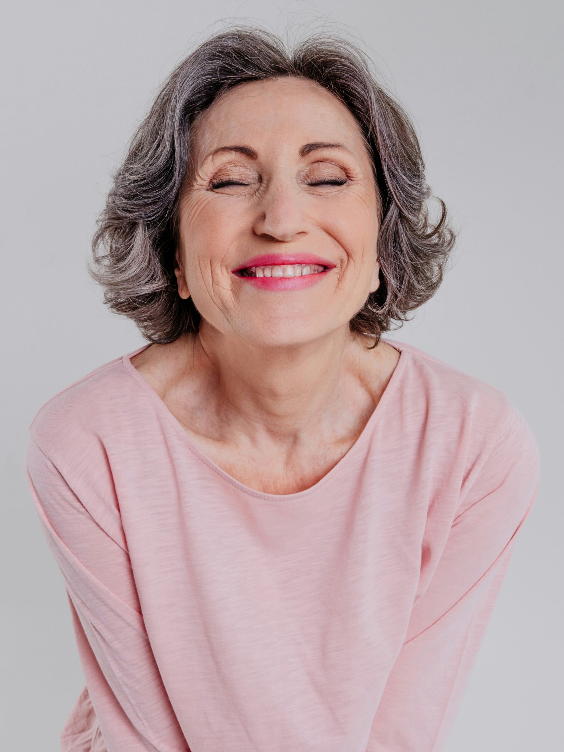 Retrato de uma mulher com cabelo curto ondulado sal e pimenta, com uma blusa rosa larga, a sorrir de olhos fechados