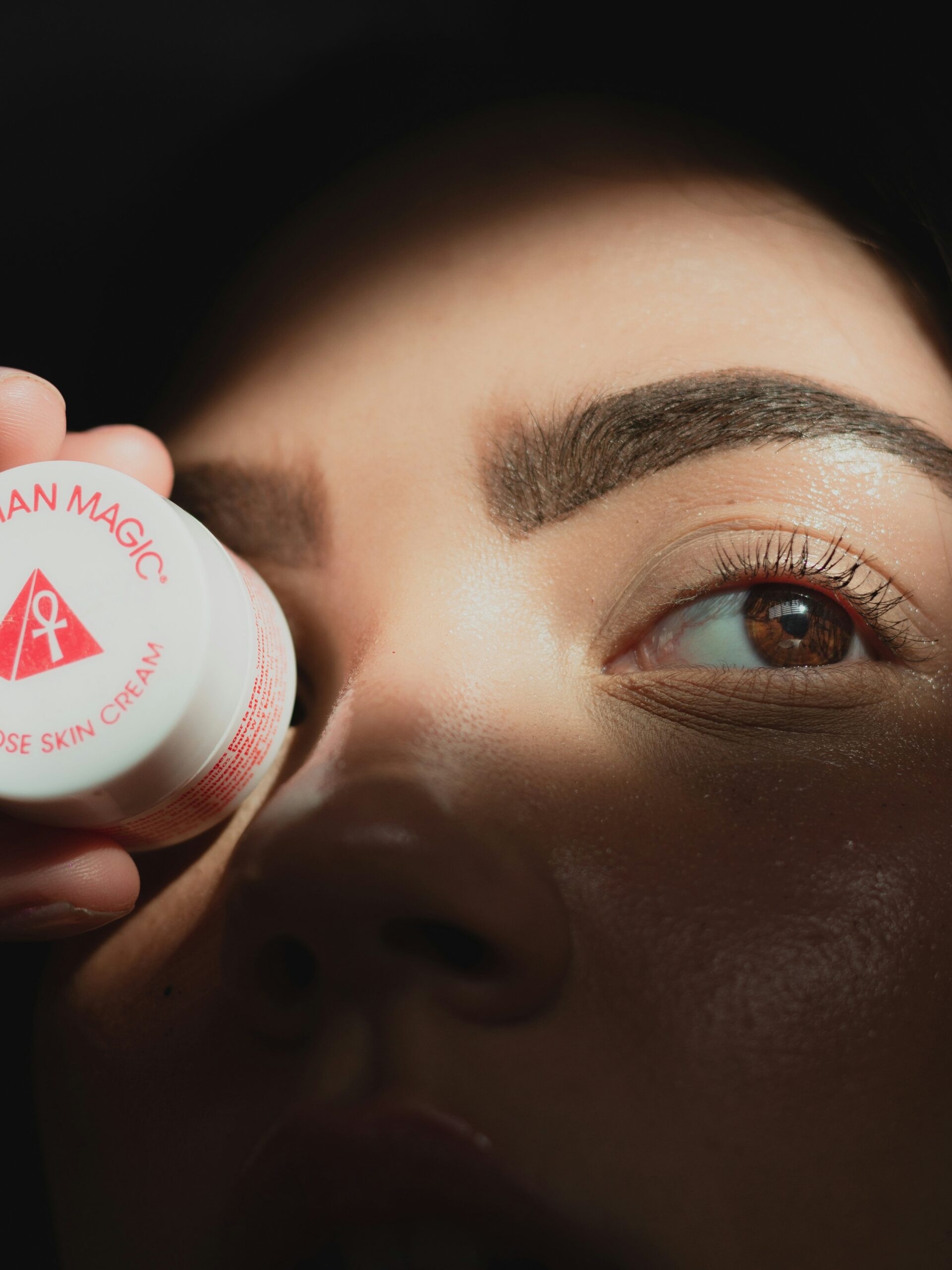Close-up do rosto de uma mulher com microblading nas sobrancelhas a segurar um pequeno boião redondo de creme em frente a um olho