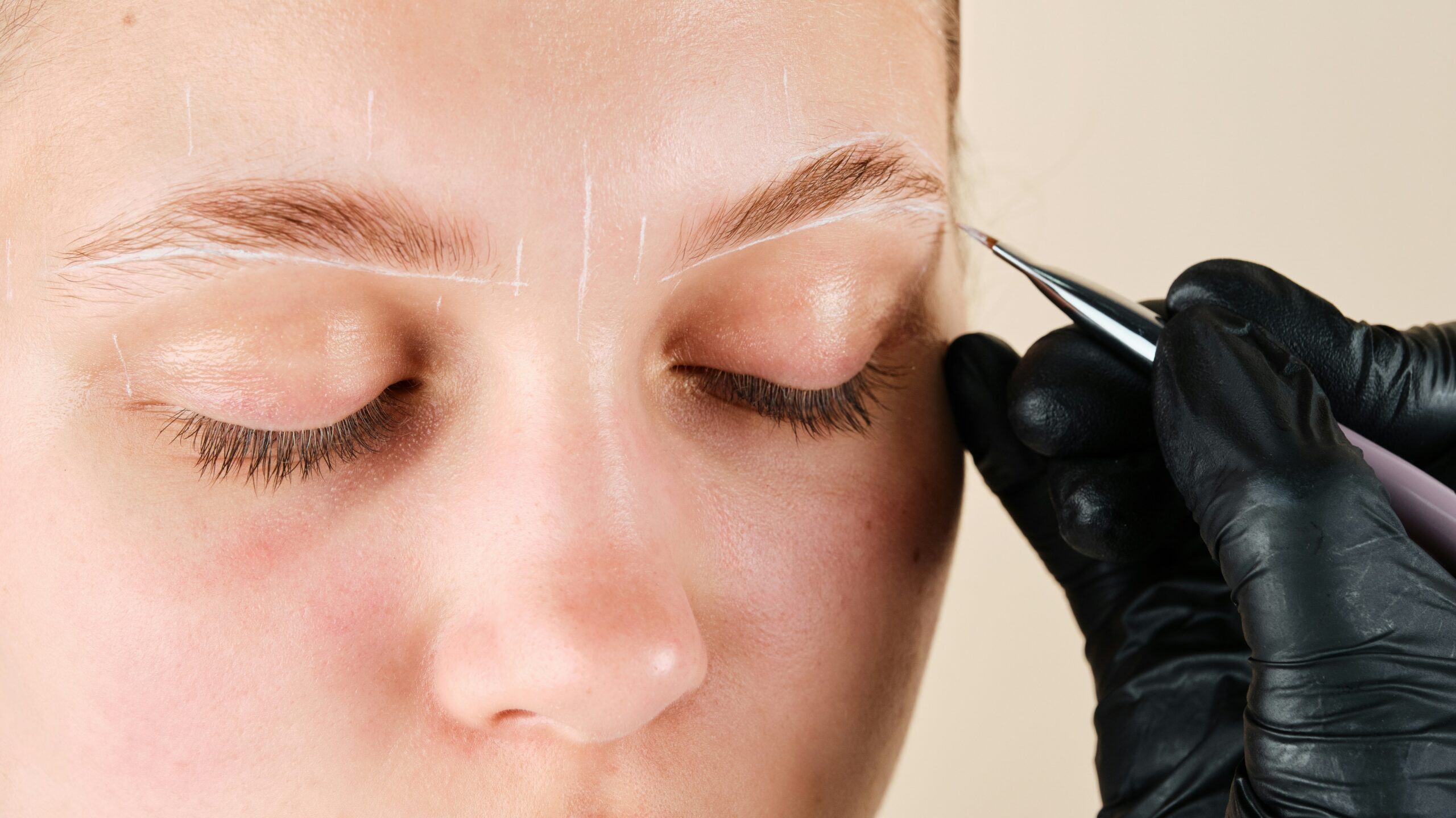 Close-up do rosto de uma mulher enquanto uma técnica desenha marcas brancas para mapear o formato desejado das sobrancelhas