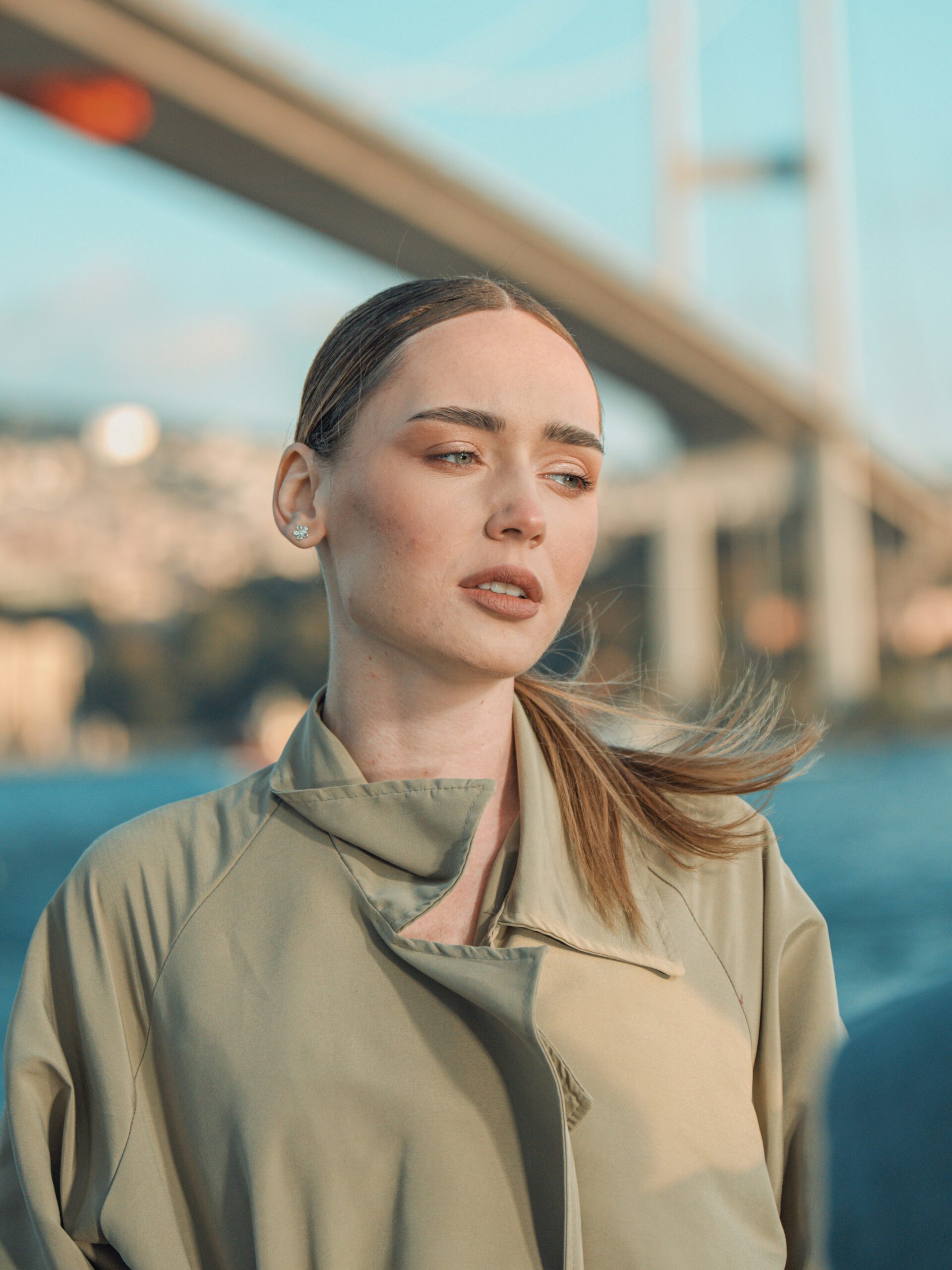Uma mulher com microblading nas sobrancelhas e cabelo claro preso, a usar um trench coat em frente a uma ponte em Istambul
