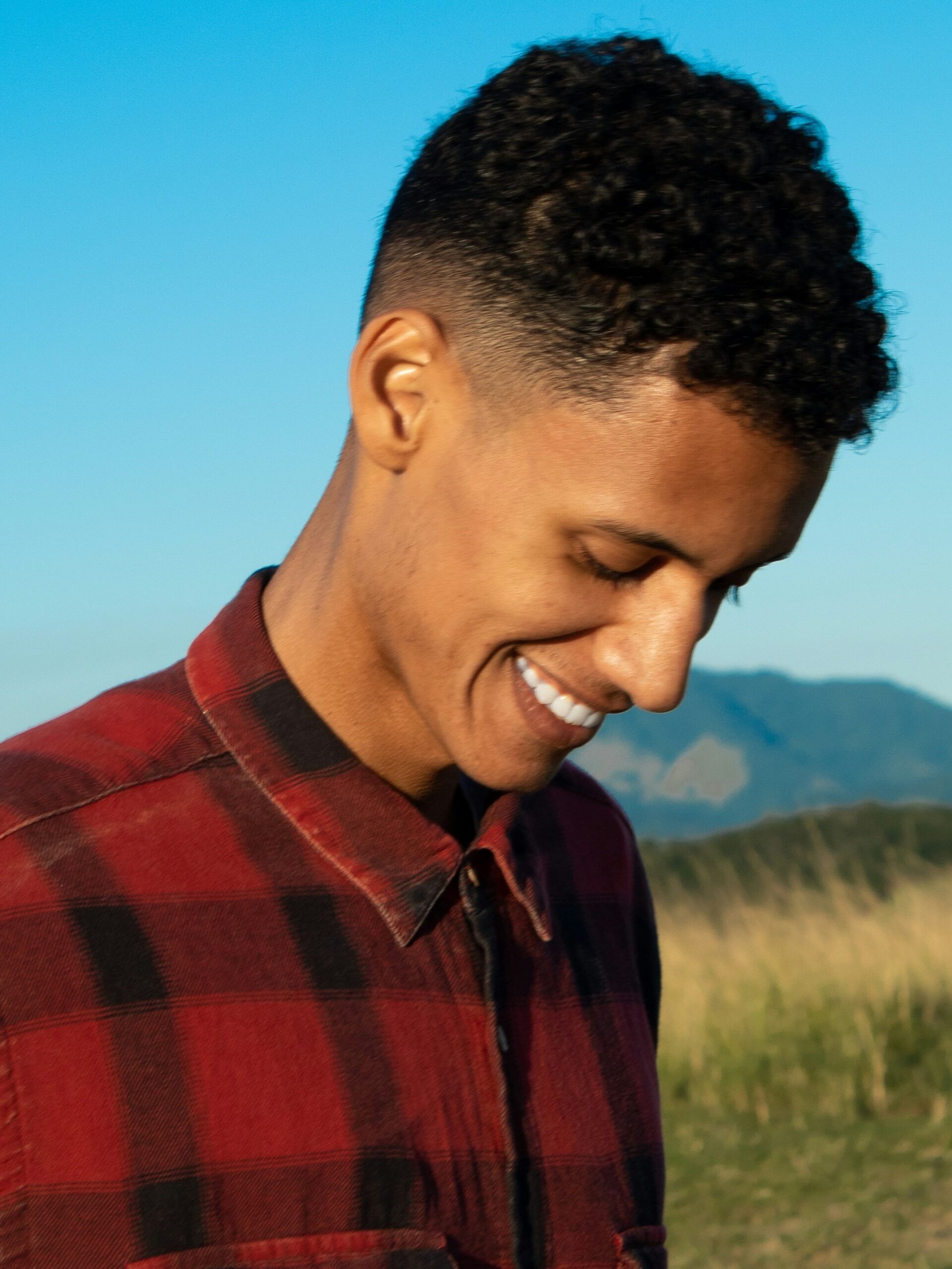 Homem a sorrir ao ar livre com montanhas e céu azul ao fundo, com cabelo encaracolado combinado com um taper fade médio