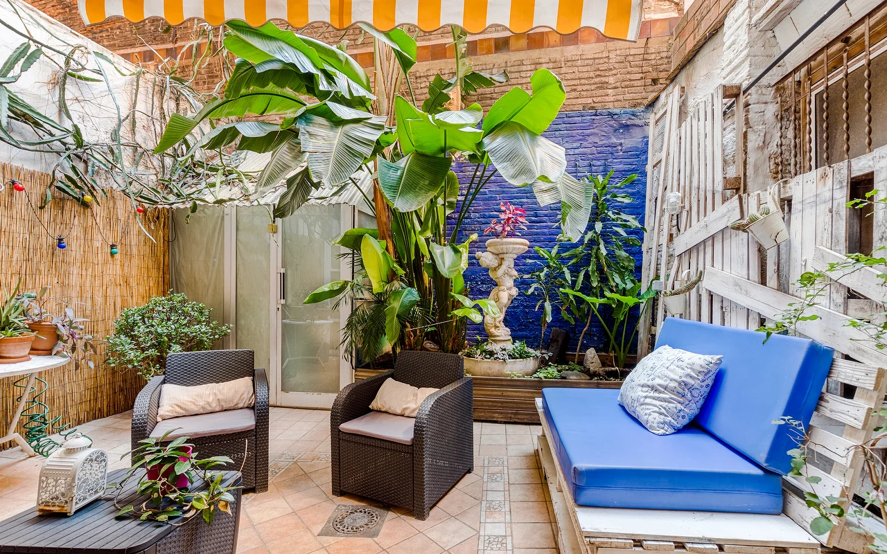 Um ptio exterior eclético com uma parede de destaque azul vibrante e plantas tropicais exuberantes. Inclui cadeires de vime escuro, um banco com almofada azul vivo e um toldo às riscas amarelas e brancas
