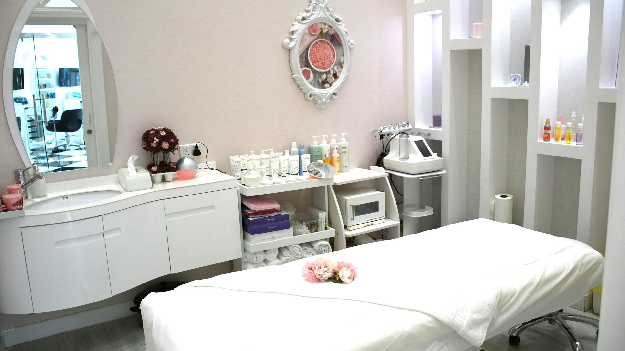 Sala de tratamento de salão luminosa, limpa e acolhedora, com marquesa, produtos nas prateleiras, espelho trabalhado e flores