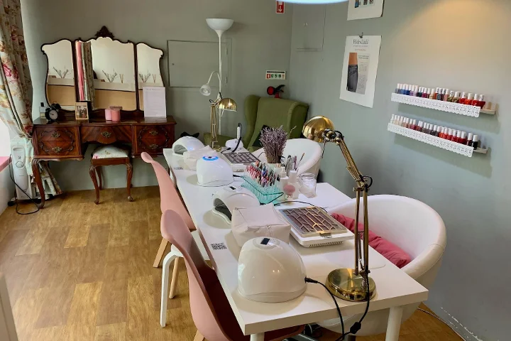 Vista interior de um salão de beleza acolhedor, especializado em manicure. Em primeiro plano, uma mesa branca comprida com lâmpadas de unhas, candeeiros de secretária e vários utensílios de manicure, ladeada por cadeiras cor-de-rosa e brancas. Ao fundo, um toucador vintage em madeira com espelho de três faces, uma poltrona verde e prateleiras brancas com filas de vernizes coloridos.
