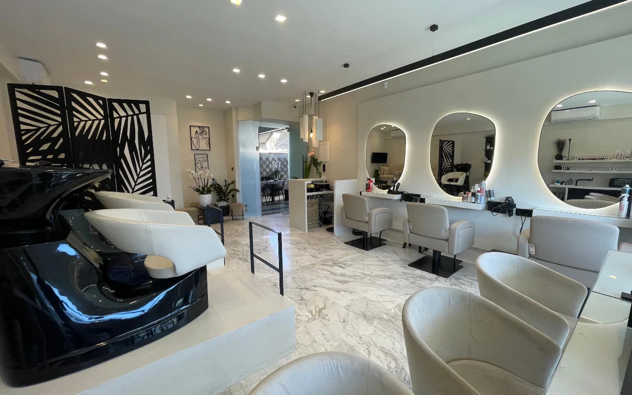 Um cabeleireiro elegante com pavimento em mármore branco, cadeiras de styling em couro creme sob espelhos ovais com contorno LED, uma estação de lavagem preta brilhante e um divisor geométrico com folhas de palmeira.