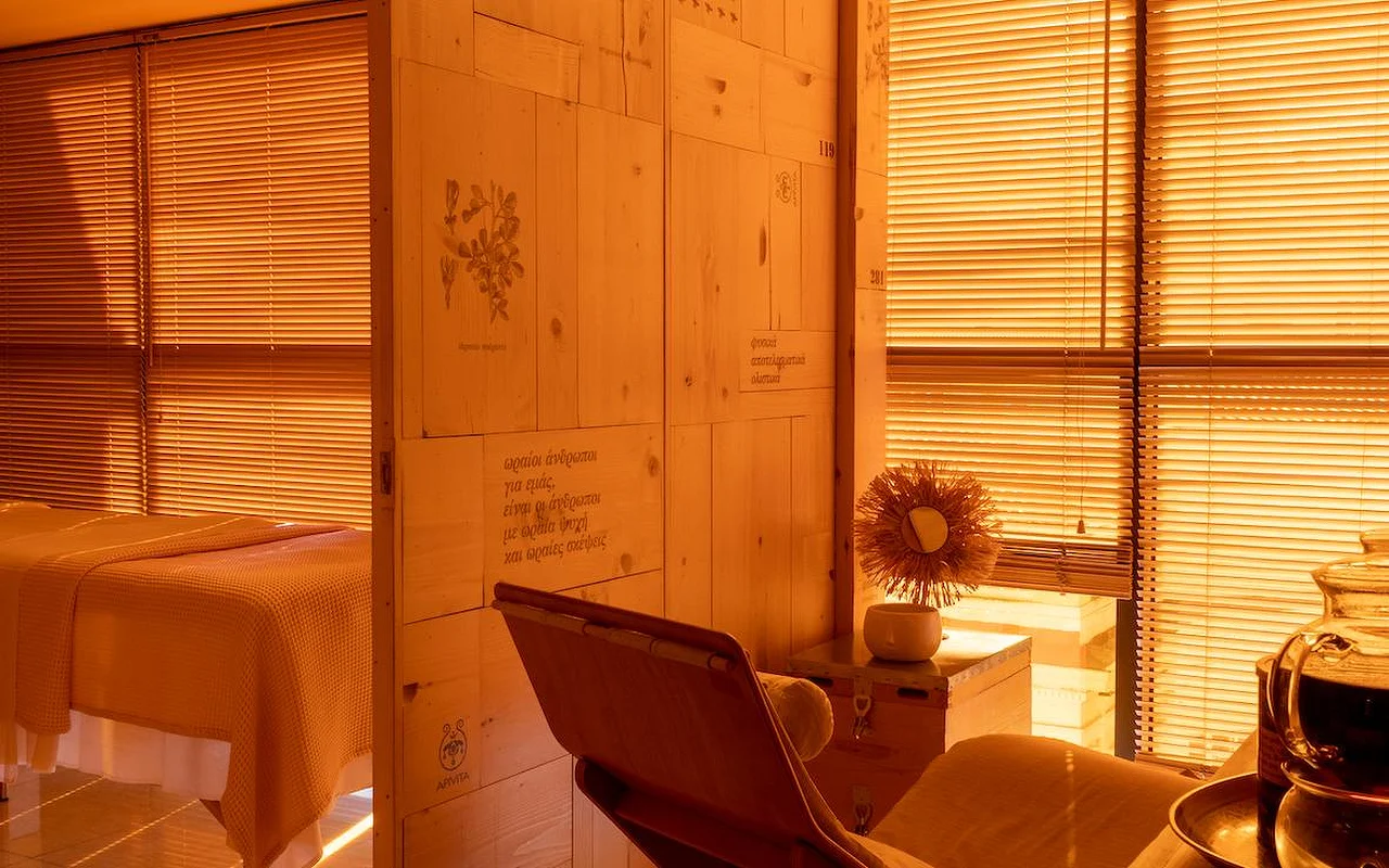 Uma sala de tratamento de spa com tons quentes de âmbar, painéis de madeira natural, ilustrações botânicas e texto grego gravados na parede, uma cama de tratamento coberta com uma manta waffle e o logótipo APIVITA no canto.