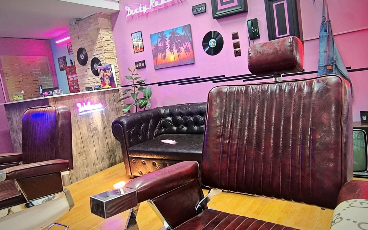Uma barbearia de estilo retro com paredes rosa e roxo, letreiros em néon, discos de vinil como decoração, uma pintura de pôr do sol com palmeiras, cadeiras de barbeiro em couro escuro e um sofá Chesterfield sobre pavimento de madeira quente.