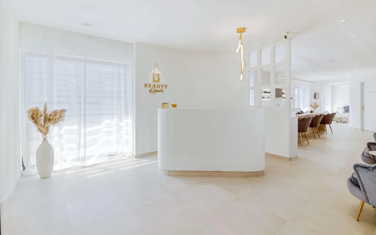 Uma receção toda em branco e luminosa com um balcão curvo, o logótipo dourado Beauty of Youth na parede, um candeeiro pendente escultórico, cortinas transparentes a filtrar a luz natural e cadeiras de tratamento em veludo castanho visíveis ao fundo.