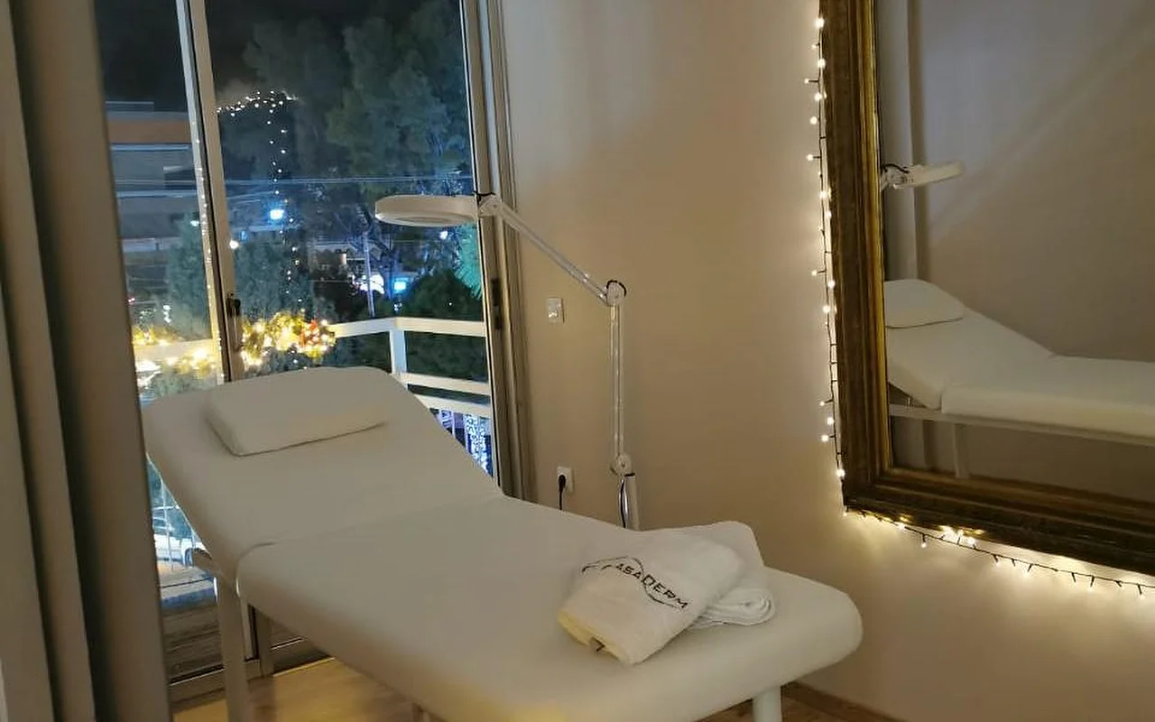 Uma sala de tratamento com iluminação suave, uma cama branca, uma toalha com a marca CasaDerm, uma lâmpada de ampliação, luzes de fada a emoldurar um espelho dourado e uma porta de varanda com vista para a paisagem noturna de Glyfada.