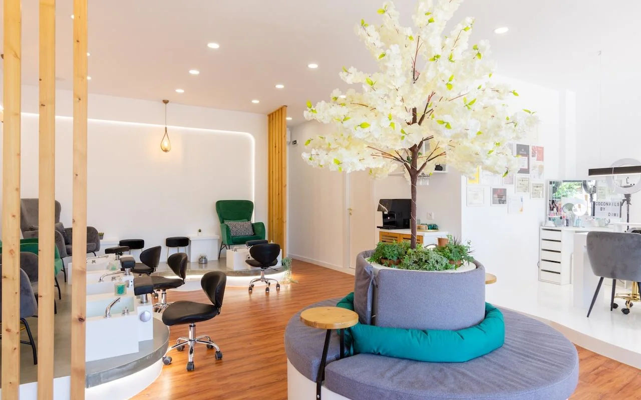 Um salão de unhas aberto e inspirado na natureza com uma grande árvore decorativa branca em flor como peça central, poltronas de veludo verde-esmeralda nas estações de pedicure, divisórias em ripas de madeira e pavimento de madeira quente.