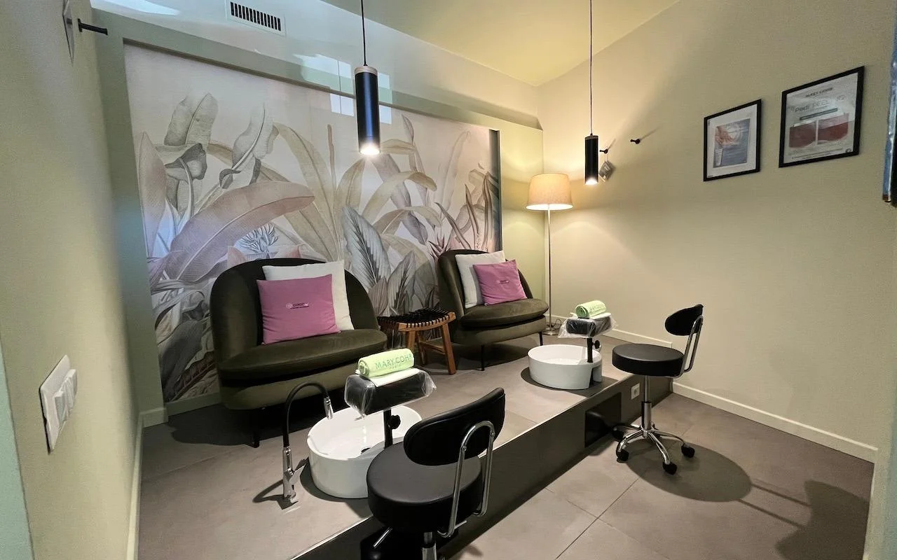 Uma estação de pedicure sofisticada com duas poltronas em veludo verde-escuro e almofadas rosa. Banhos de pés brancos estão embutidos numa plataforma cinzenta elevada, com um mural de grandes folhas tropicais na parede traseira.
