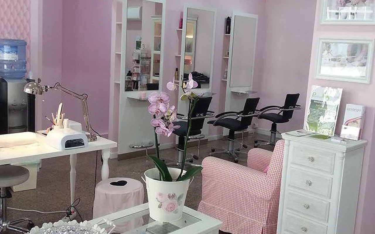 Interior de um salão de cabelo e unhas luminoso, com tema cor-de-rosa. À esquerda, uma estação de manicure branca equipada com candeeiro de secretária e aparelho de secagem de unhas. Ao centro, uma orquídea num vaso branco sobre uma mesa de café junto a uma poltrona rosa e branca aos quadrados. Ao fundo, várias estações de estilismo com espelhos altos e cadeiras pretas de salão contra paredes rosa-claro.