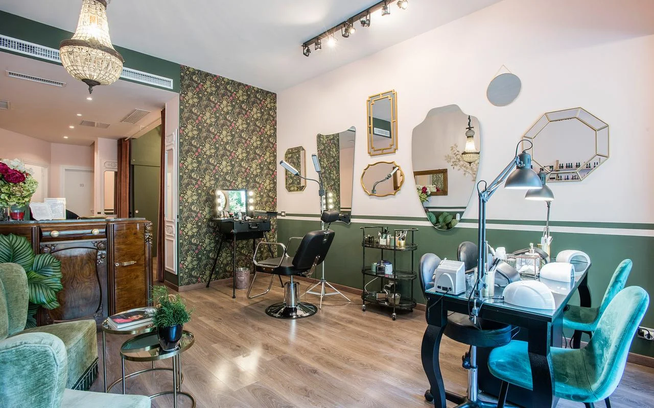 Um salão vintage-chic com paredes brancas e verde-floresta. Apresenta papel de parede com padrão floral, um conjunto de espelhos com molduras douradas de formas irregulares, uma estação de maquilhagem profissional e assentos em veludo verde.