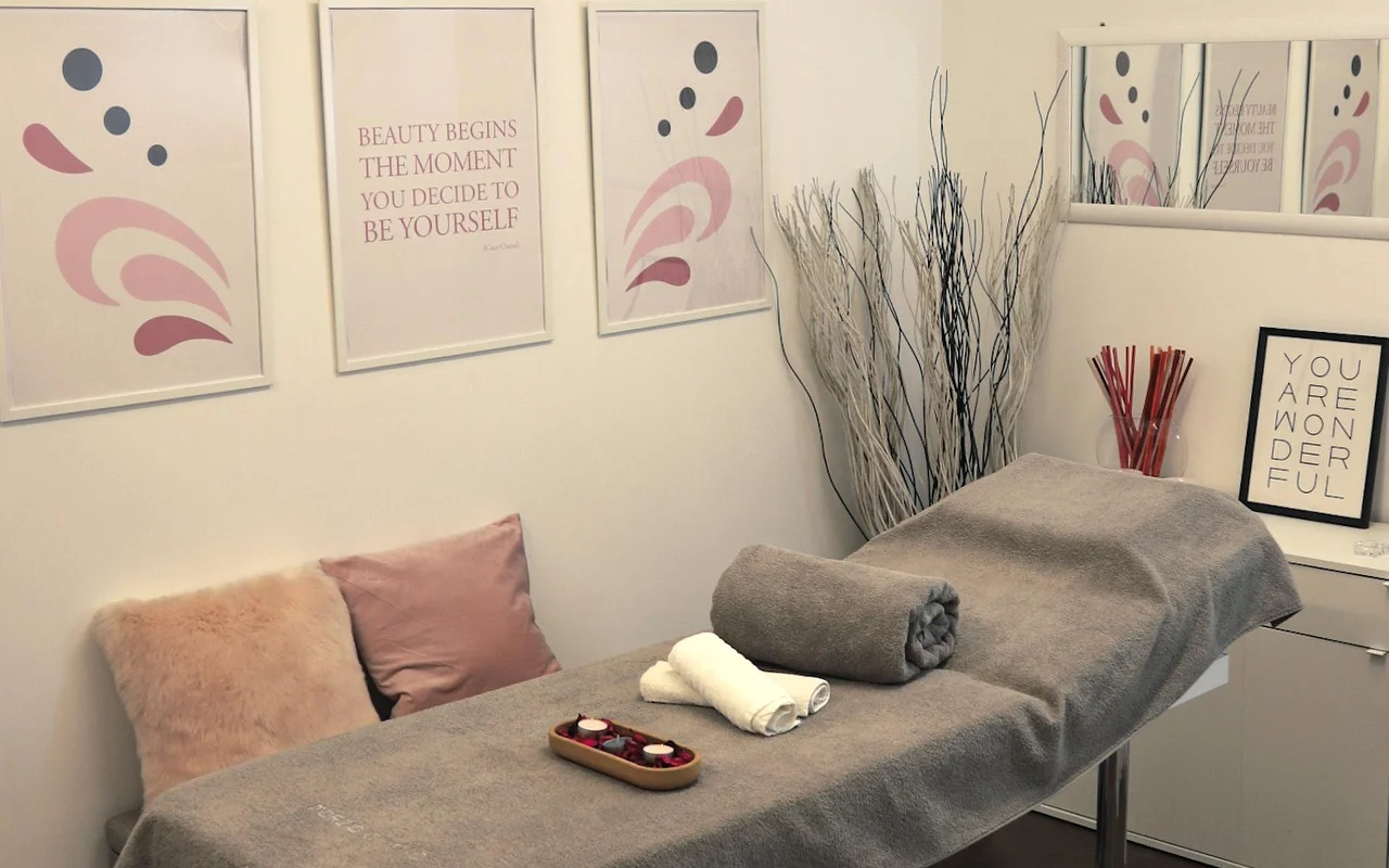 Uma sala de tratamento luminosa com mesa de massagem coberta por uma toalha cinzenta. A parede está decorada com arte abstrata em tons pastel e frases motivacionais, enquanto ramos brancos decorativos e velas criam uma atmosfera calma.