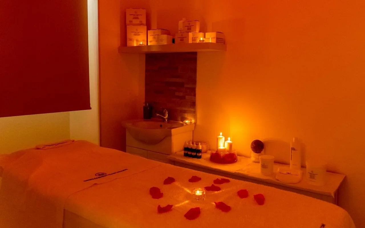 Uma sala de massagem com iluminação quente em tons de laranja e velas a cintilar. Pétalas de rosa vermelha estão dispostas em padrão circular à volta de uma vela central na mesa de tratamento, com produtos de beleza organizados numa prateleira atrás.