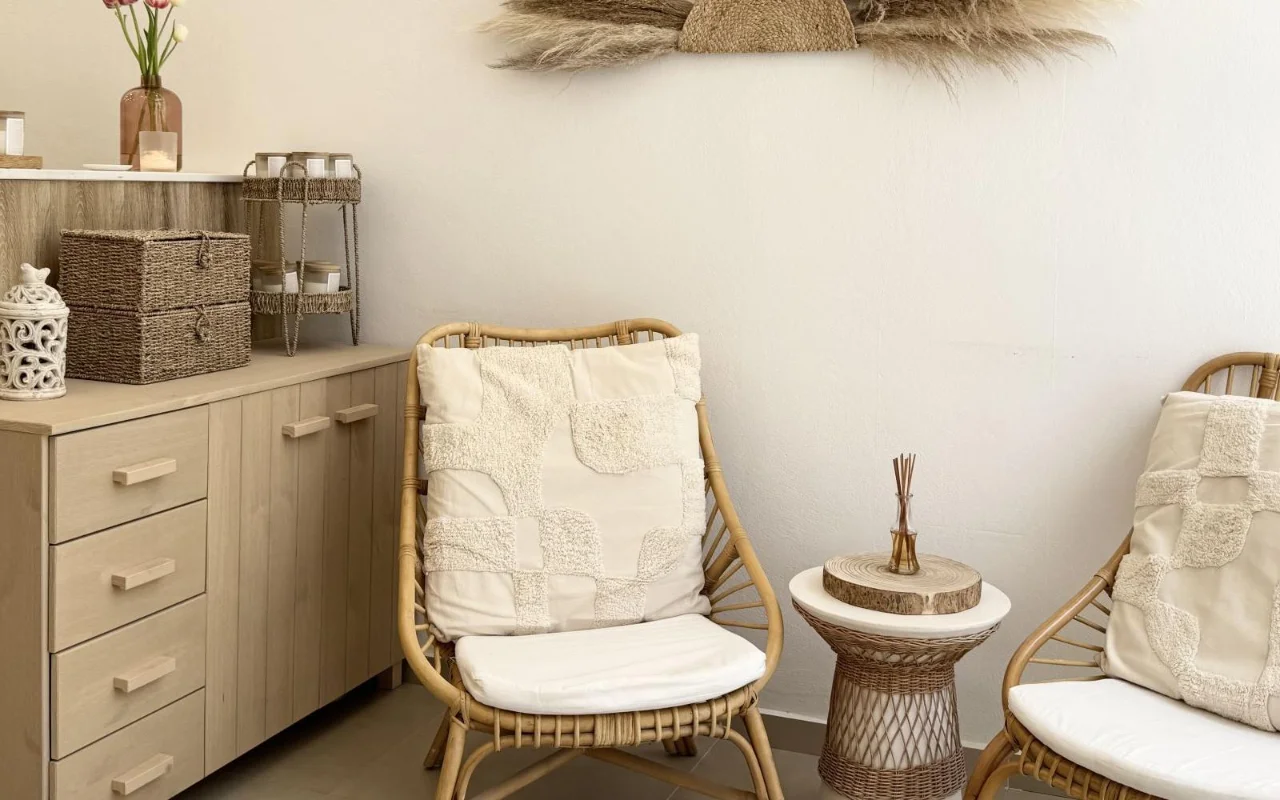 Uma sala de espera tranquila com poltronas de rattan, almofadas em tons creme com textura, uma mesa de apoio de vime com difusor, cestos de arrumação em fibra natural e erva dos pampas seca na parede acima de armários de madeira clara.