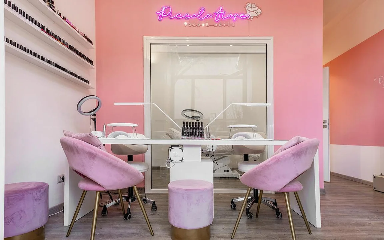 Uma estação de manicure com paredes cor-de-rosa vibrante e um letreiro néon rosa choque. Cadeiras em veludo lilás com pernas douradas estão junto a uma secretária branca minimalista equipada com lâmpadas LED profissionais para unhas.