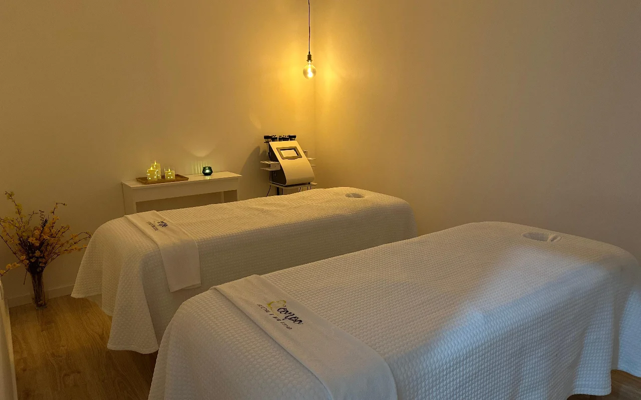 Uma sala de massagem ou tratamento tranquila com duas marquesas lado a lado, cobertas com toalhas e lençóis brancos impecáveis. A sala é iluminada por uma única lâmpada quente suspensa e pequenas velas numa mesa de apoio, criando uma atmosfera serena. Uma máquina cosmética especializada está ao fundo, entre as marquesas.