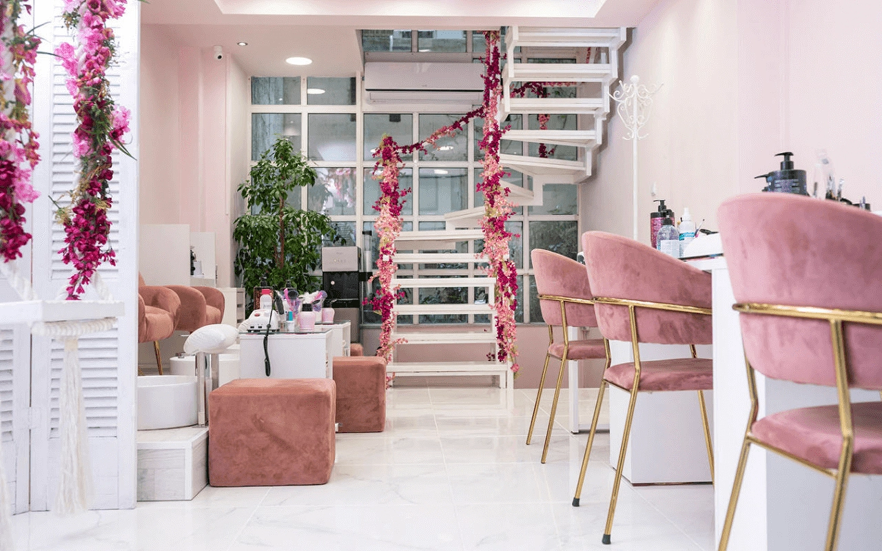 Um salão de unhas feminino com cadeiras em veludo rosa-velho, uma escadaria em espiral branca decorada com flores artificiais cor-de-rosa, divisórias com portadas, e estações de manicure com pés dourados sobre pavimento com efeito de mármore.