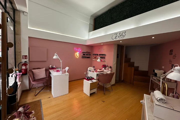 Um estúdio de unhas elegante com paredes rosa-claro e chão de madeira quente. A sala tem várias secretárias de manicure brancas combinadas com cadeiras de veludo rosa-velho. Um letreiro néon cor-de-rosa em forma de mão com um frasco de verniz e a palavra 'Nails' brilha na parede, e filas de vernizes estão expostas em prateleiras pequenas. Um mezanino com uma parede de sebe artificial verde é visível acima.