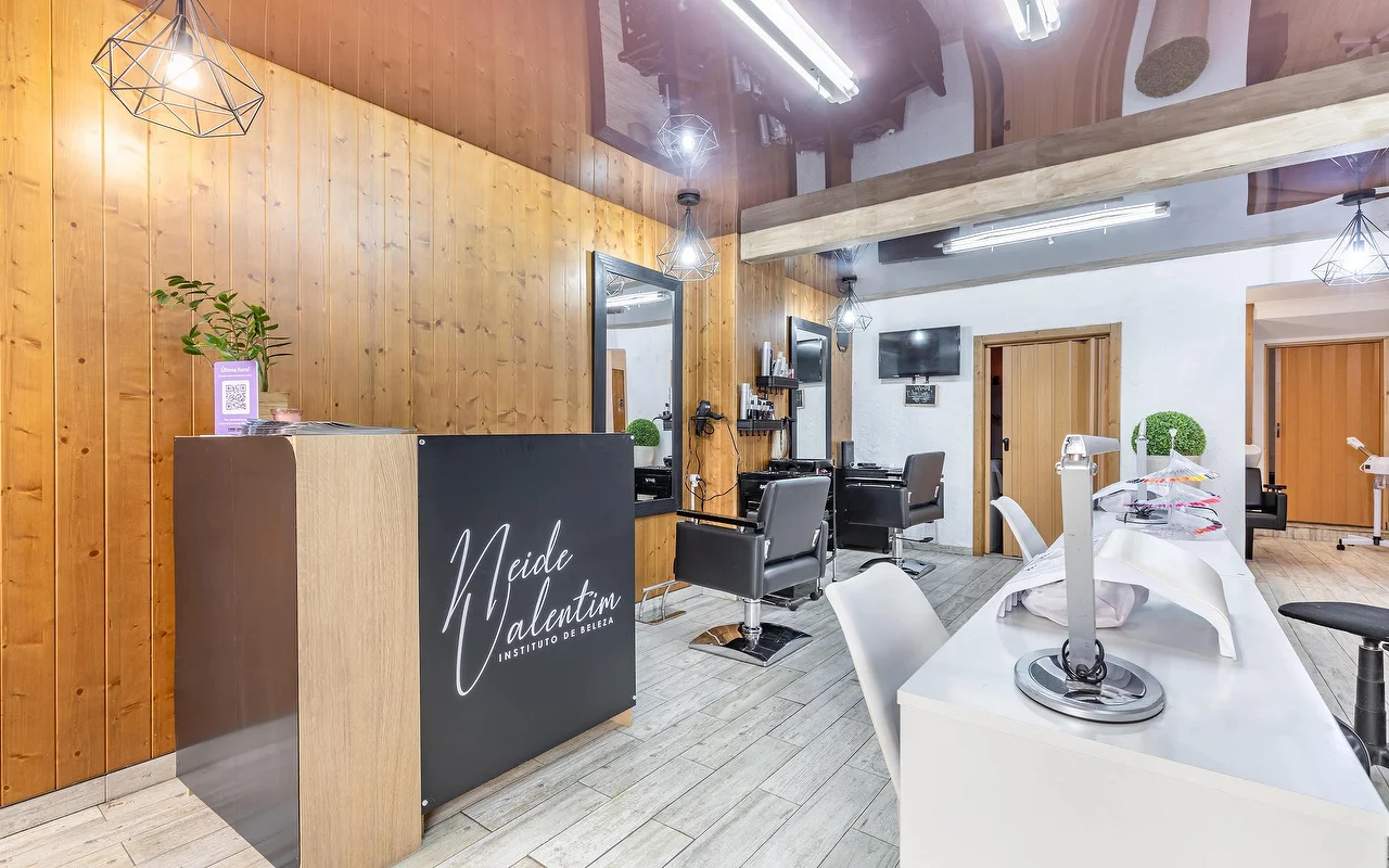 Um salão de beleza contemporâneo com uma receção à esquerda com a marca 'Neide Valentim Instituto de Beleza'. As paredes têm revestimento vertical em madeira quente e espelhos grandes. Cadeiras de estilismo pretas estão em frente aos espelhos, enquanto uma estação de manicure branca com cadeiras brancas se estende ao longo do lado direito do chão de azulejo cinzento.