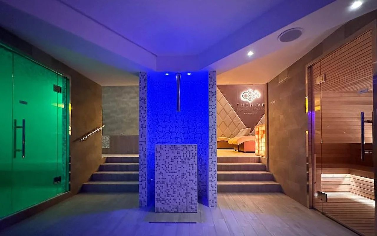 Área termal de um spa de luxo com iluminação atmosférica em azul e verde. O espaço inclui uma sauna finlandesa em madeira, entrada para banho turco e um elemento central em mosaico com degraus que conduzem a zonas de relaxamento.