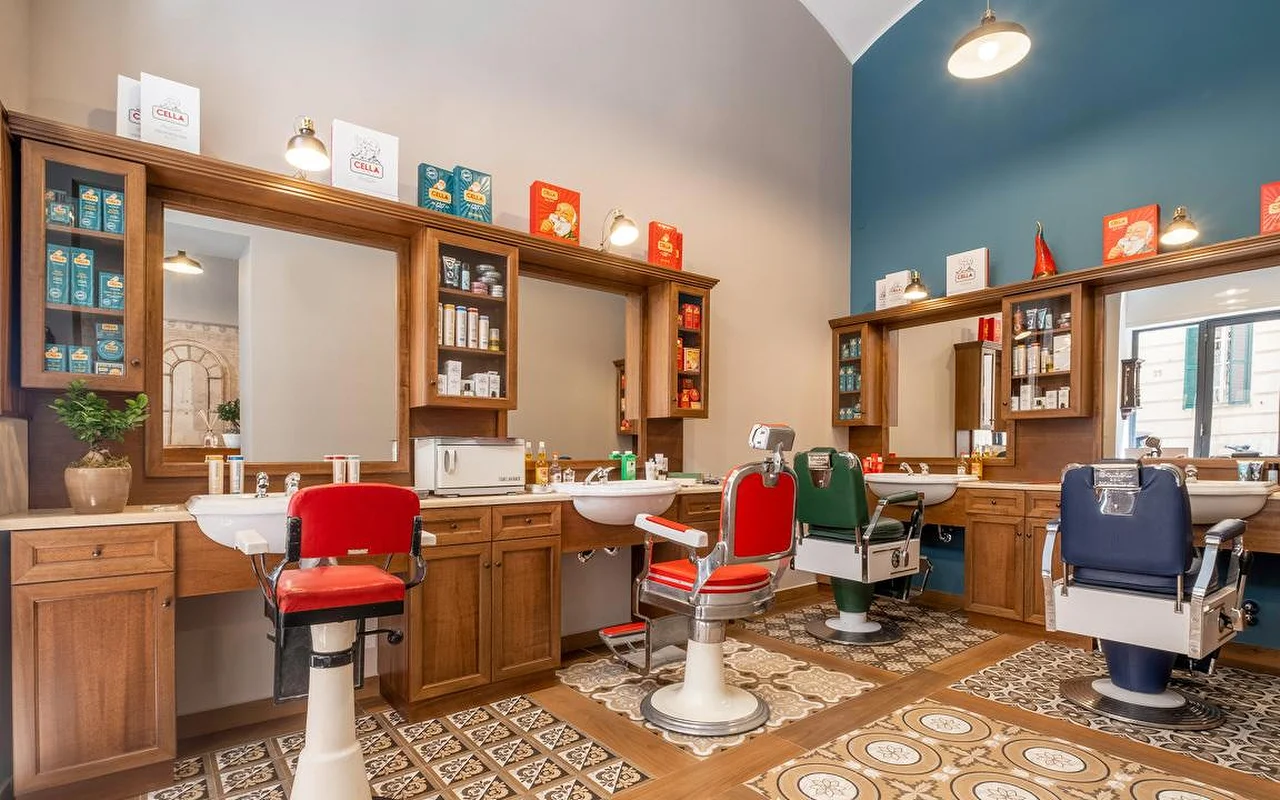Uma barbearia tradicional de alta qualidade com armários de madeira ricamente trabalhados, azulejos de chão com padrão vintage e cadeiras de barbeiro profissionais em vermelho, verde e azul-marinho. Produtos de grooming tradicionais estão expostos nas estantes.