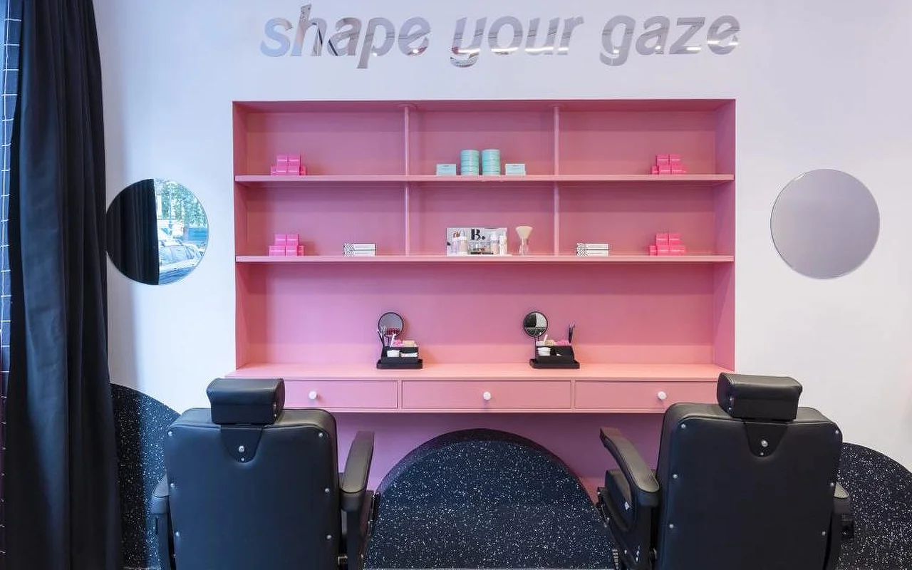 Um estúdio especializado em sobrancelhas e pestanas com uma estante cor-de-rosa viva a exibir produtos de beleza, letras prateadas 'shape your gaze' na parede e cadeiras de tratamento em couro preto sobre um chão escuro salpicado.