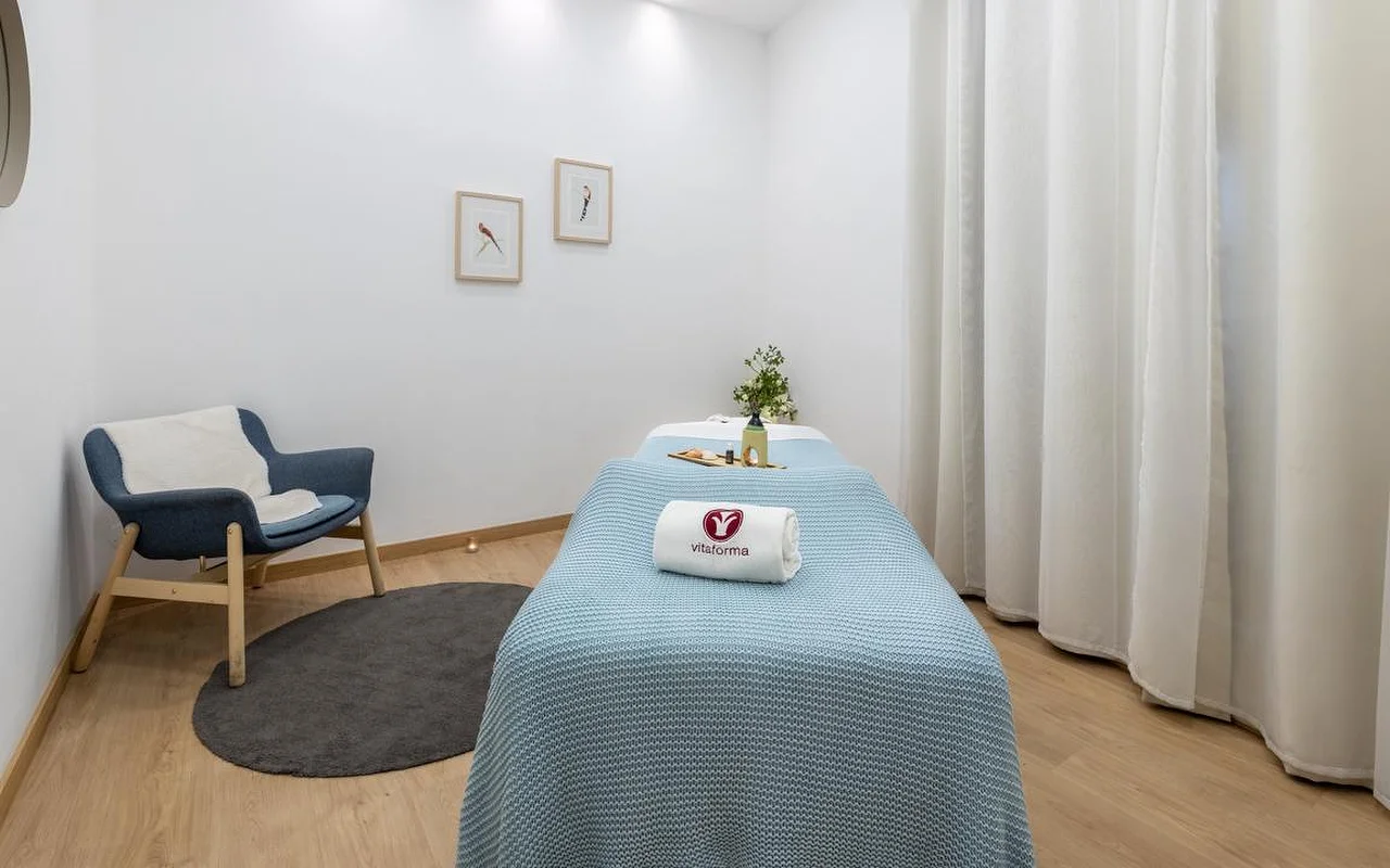 Uma sala de spa limpa e minimalista com paredes brancas e chão de madeira clara. Uma marquesa de tratamento está arrumada com uma manta azul-clara de malha e uma toalha branca enrolada com o logótipo 'Vitaforma'. No canto, uma poltrona azul elegante assenta sobre um tapete redondo cinzento, e cortinas brancas compridas pendem junto à janela.