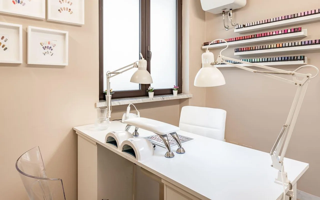 Uma estação de unhas dedicada no AG The Beauty Room's, com uma secretária branca e limpa, dois candeeiros UV, luzes de trabalho ajustáveis e prateleiras a exibir filas de vernizes coloridos. Desenhos de nail art emoldurados decoram as paredes em tons quentes.