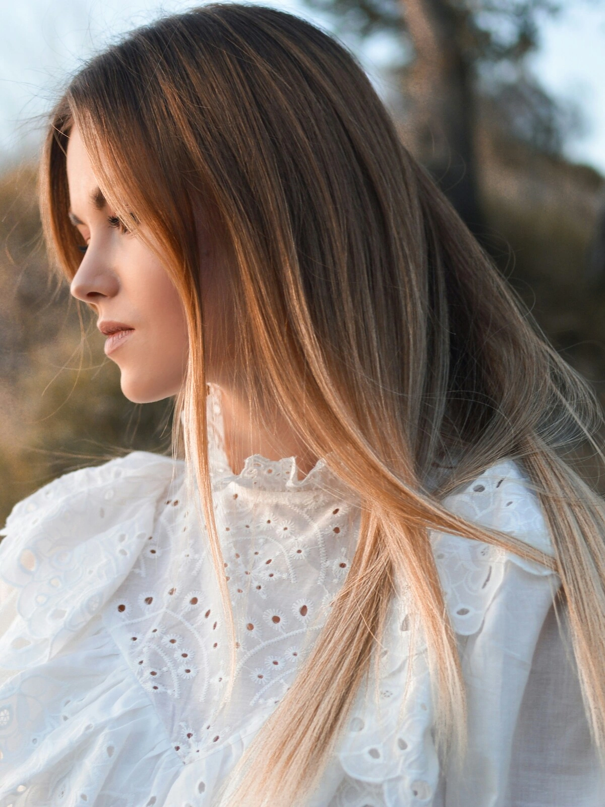 Vista de lado de uma mulher ao ar livre num ambiente natural, com balayage sobre cabelo liso em que o castanho se esbate em tons loiros