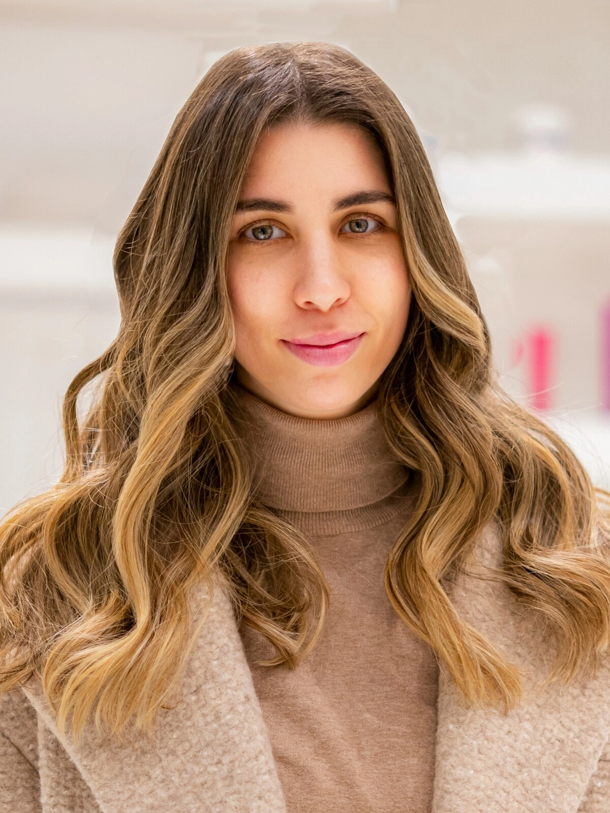 Uma mulher com uma camisola de gola alta e um casaco de lã cor de camelo, com um balayage suave e ondulado em que o castanho se esbate em tons caramelo