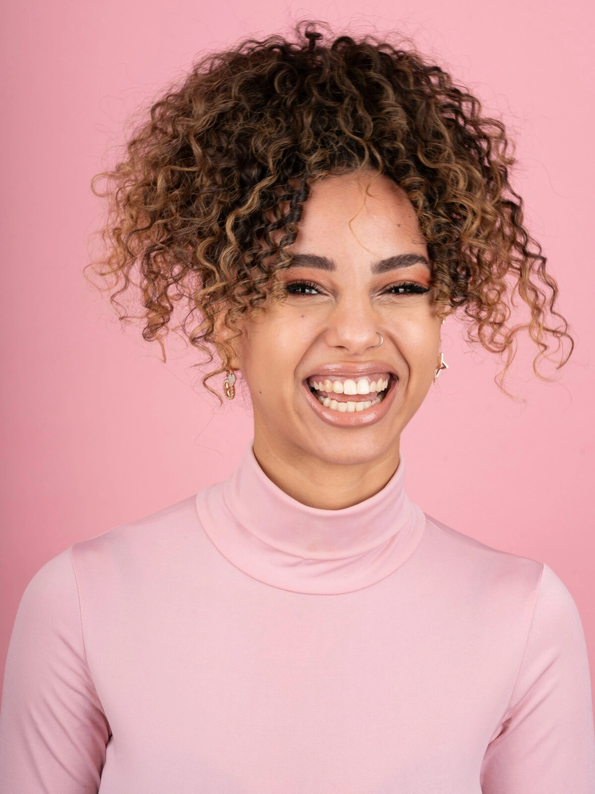 Foto de estúdio de uma mulher sorridente com balayage sobre cabelo afro, vestida com uma camisola de gola alta cor-de-rosa suave