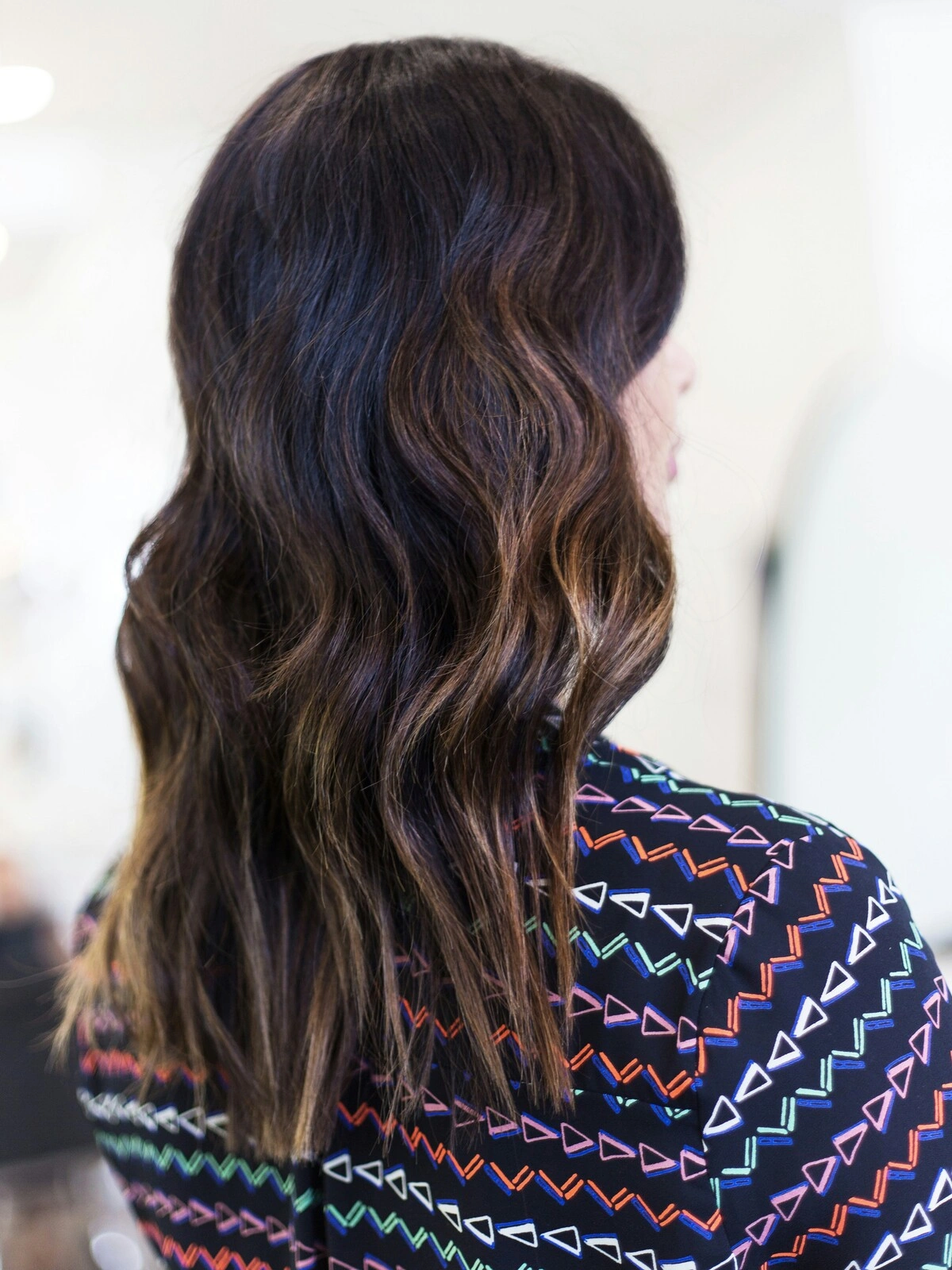 Vista de trás da cabeça de uma mulher com balayage em ondas suaves, em que o cabelo preto se esbate em tons castanhos quentes